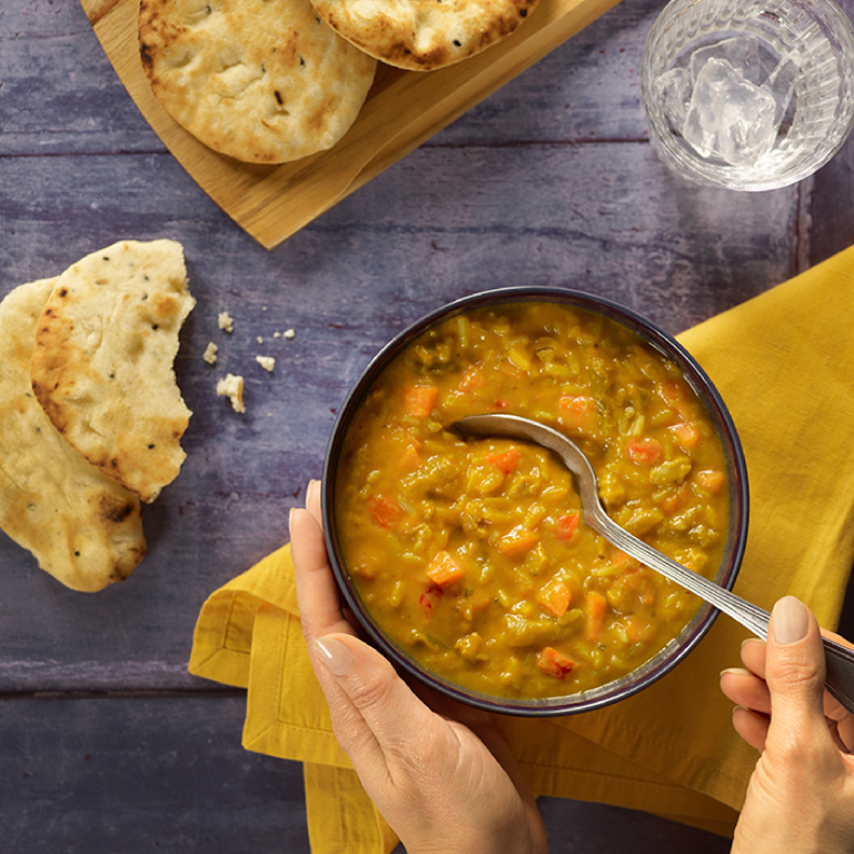 Our Jaldee Eats Moroccan Chicken Soup in a bowl, with a spoon inserted and hands shown to be stirring it up. Surrounding the bowl is some tasty naan bread, a napkin and water. Our halal meals make dinner quick and easy.